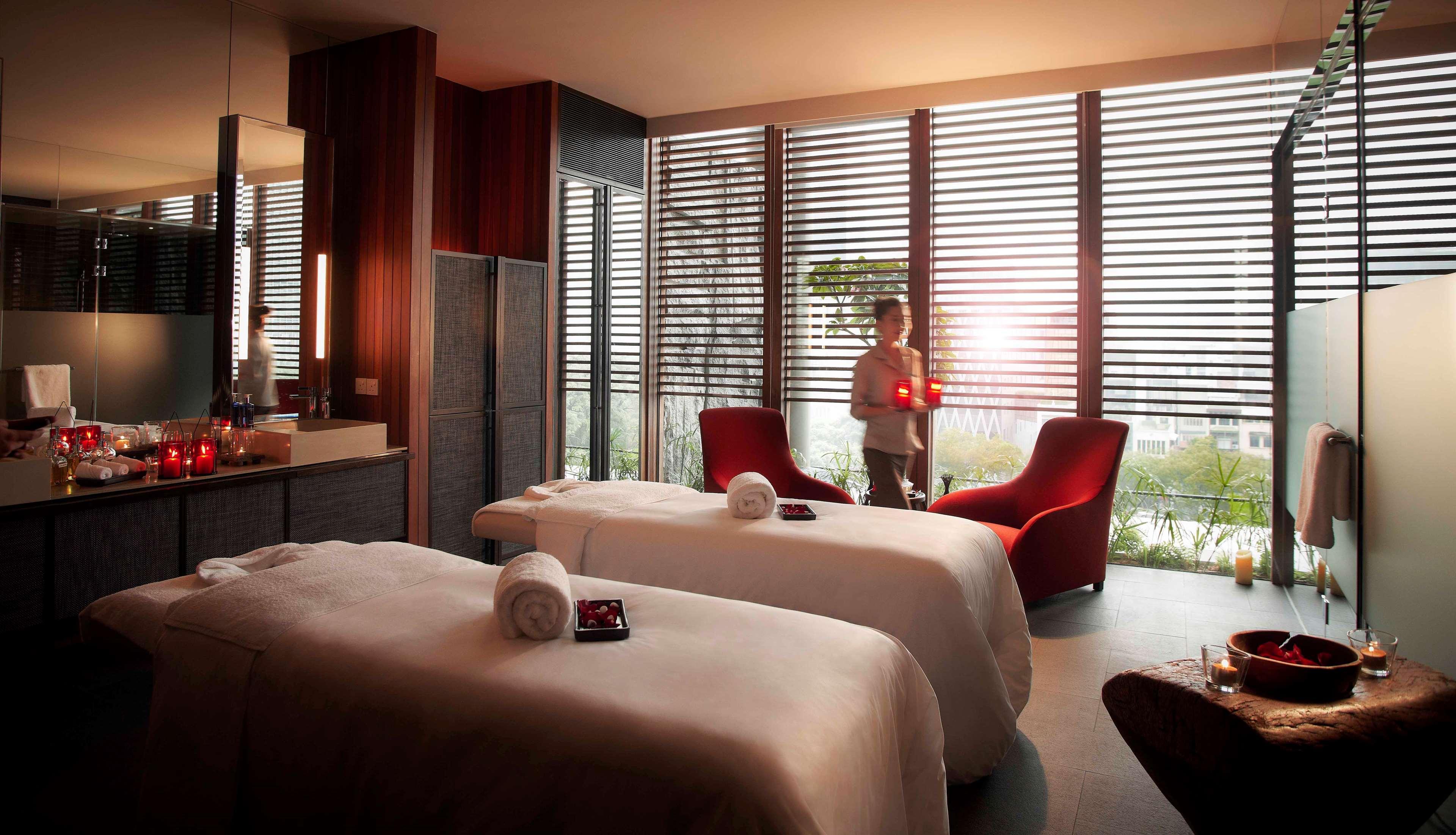 Parkroyal Collection Pickering, Szingapúr Kültér fotó The photo shows a tranquil spa room with a calming ambiance. There are two massage tables with white linens and neatly rolled towels placed on them. In the background, large windows with horizontal slats allow soft light to filter in, creating a sere