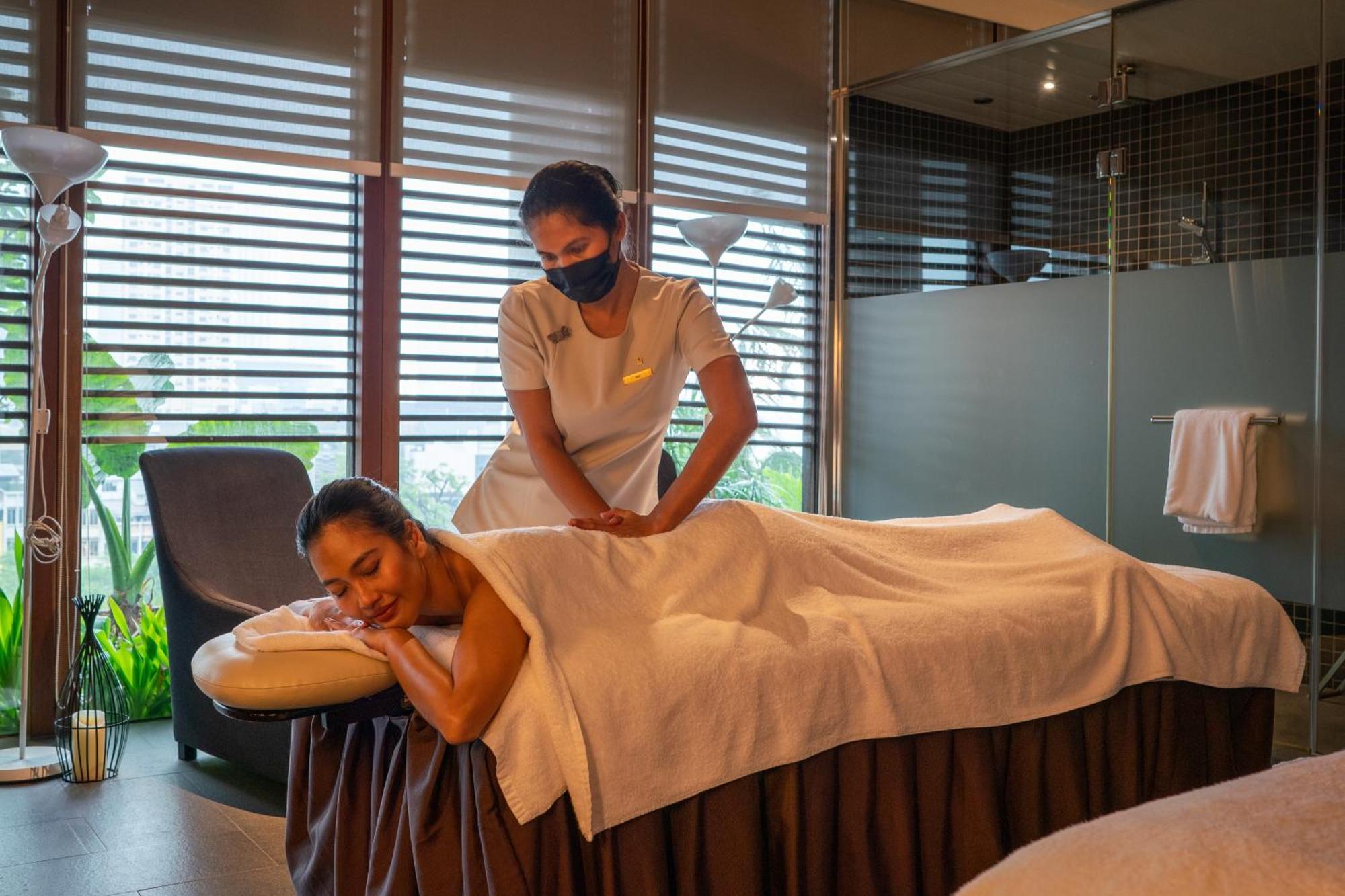Parkroyal Collection Pickering, Szingapúr Kültér fotó The photo shows a serene spa setting where a therapist is giving a massage to a client. The client is lying face down on a massage table, covered with a towel, and appears to be relaxed. The therapist is positioned at the client's back, providing the