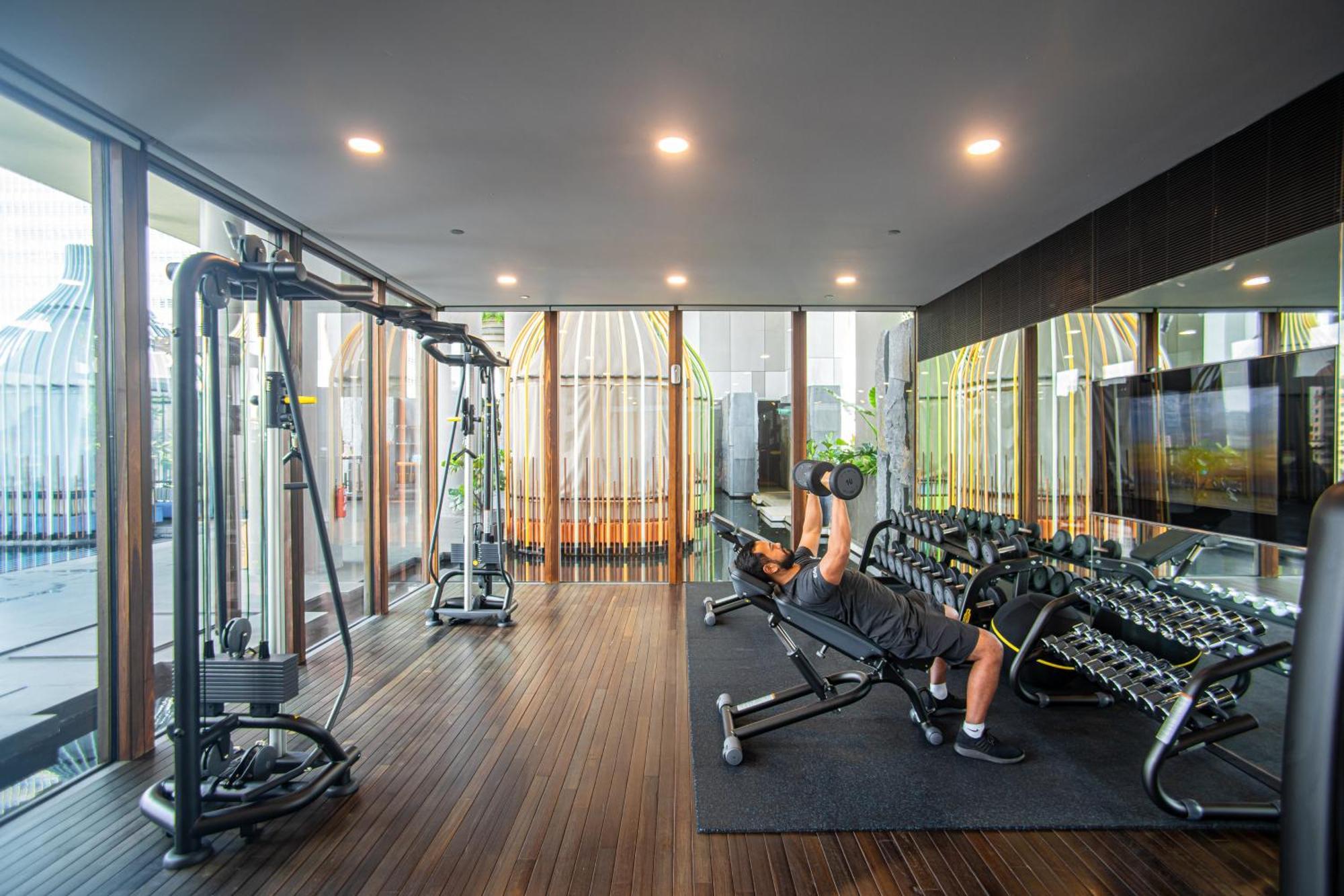 Parkroyal Collection Pickering, Szingapúr Kültér fotó The photo shows a modern gym or fitness area that features various exercise equipment. In the foreground, a person is lying on a bench and performing a dumbbell press with weights. Surrounding them are several dumbbells neatly arranged on a rack. The