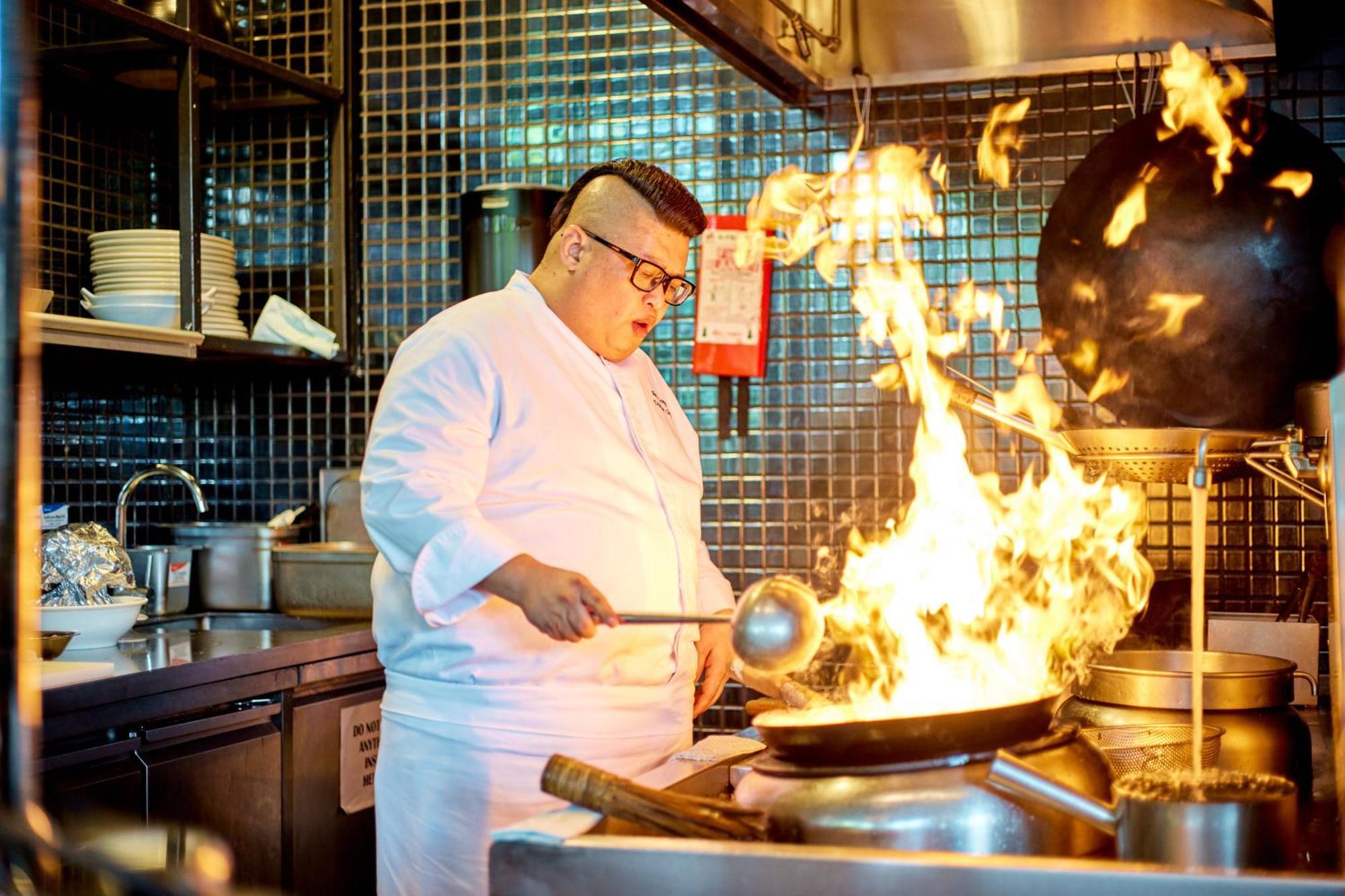 Parkroyal Collection Pickering, Szingapúr Kültér fotó The photo shows a chef in a kitchen, wearing a white chef's jacket and glasses. He is actively cooking in front of a large wok that is producing a dramatic surge of flames. The kitchen has a modern look, with tiled walls and a variety of kitchen equi