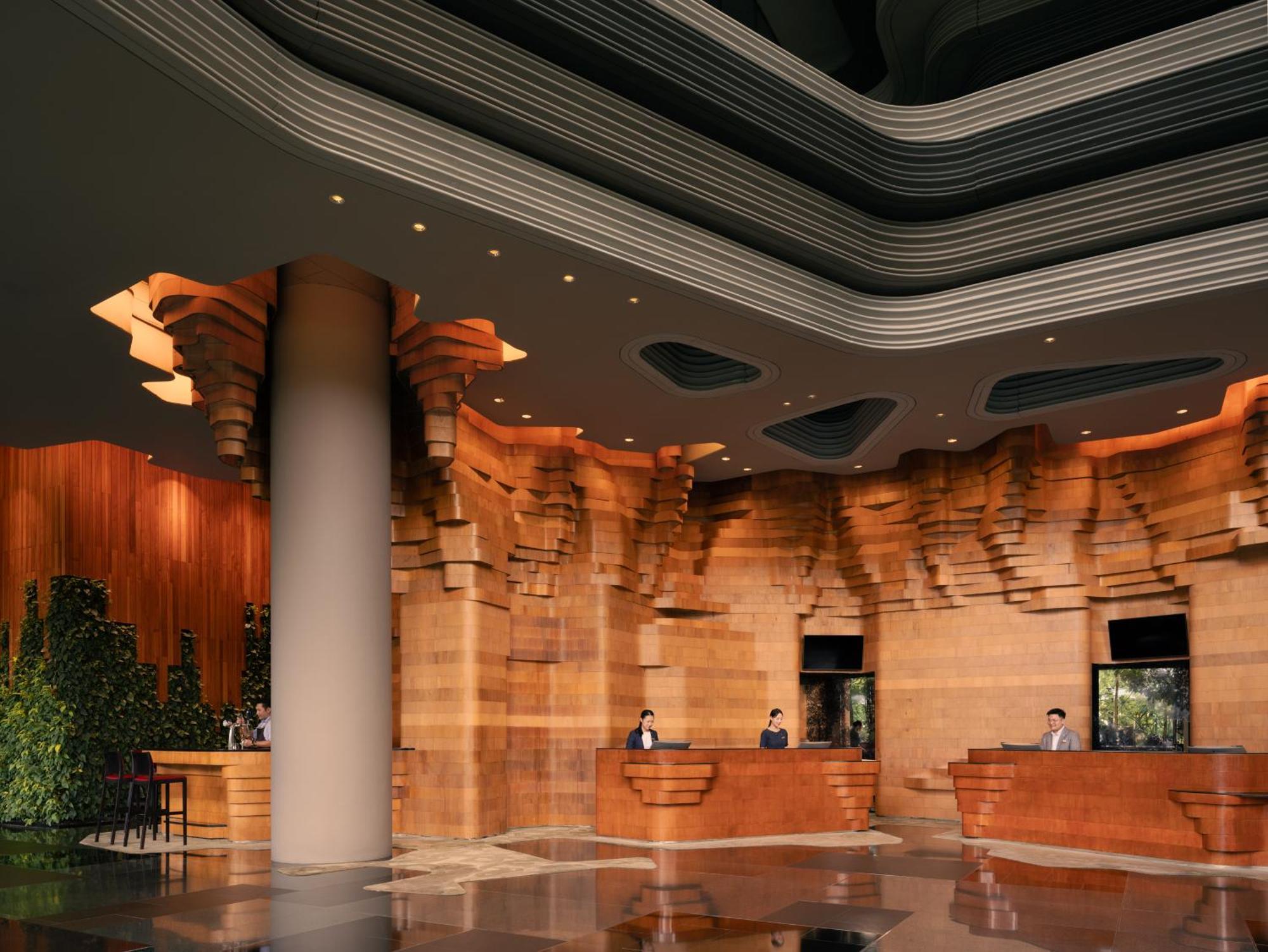 Parkroyal Collection Pickering, Szingapúr Kültér fotó The image shows a modern hotel or building lobby with a sophisticated design. The interior features richly textured wooden walls that resemble natural formations, creating a warm and inviting atmosphere. The reception area has several service counter