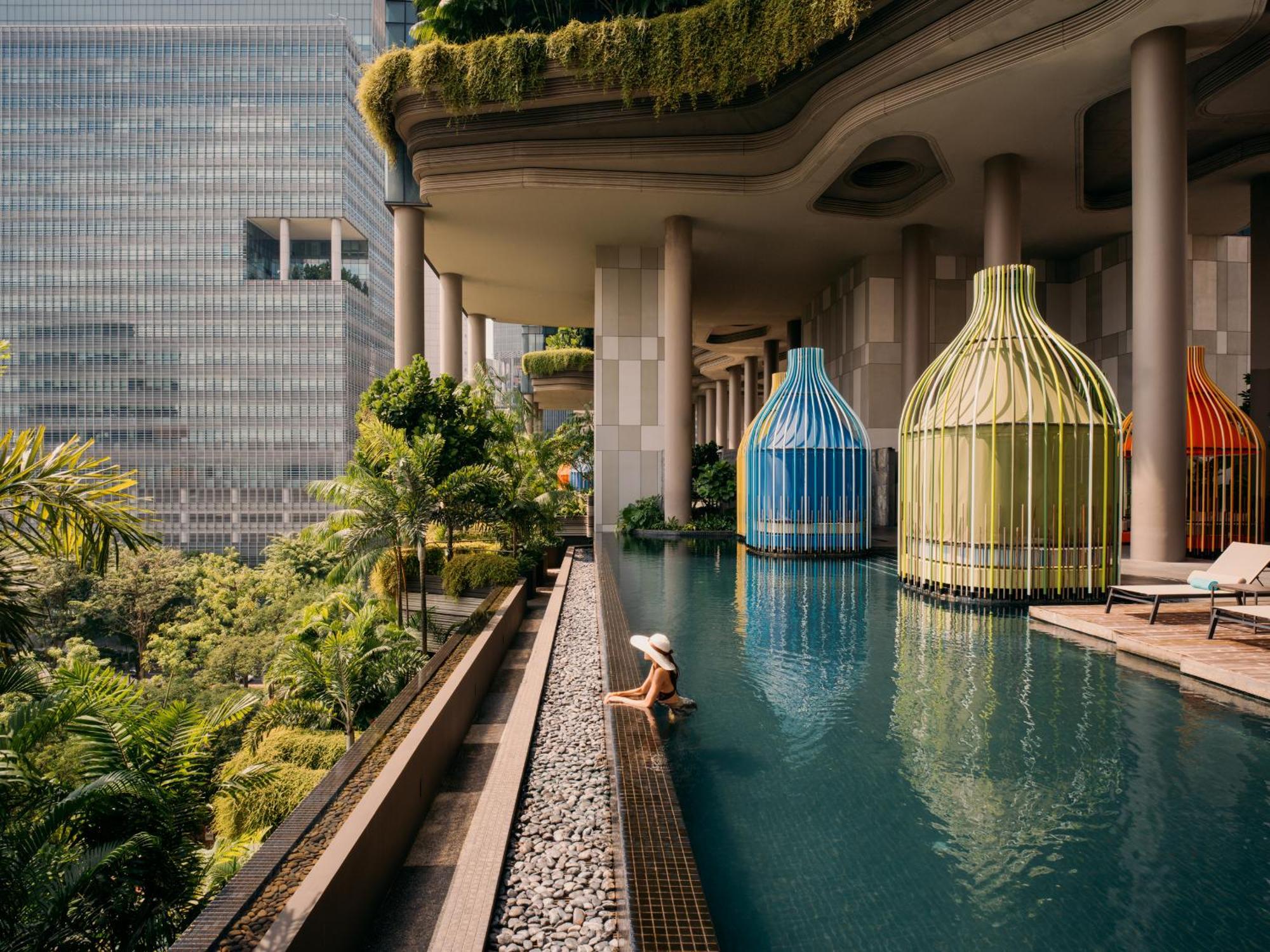 Parkroyal Collection Pickering, Szingapúr Kültér fotó The photo depicts a beautiful, modern swimming pool area surrounded by lush greenery. There are distinctive, colorful structures resembling large birdcages in blue and green, which add an artistic touch to the scene. A person is seen lounging peacefu